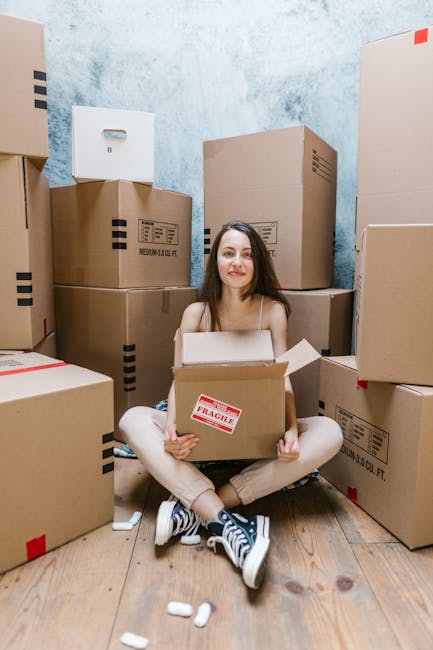 A person using masking tape to securely seal a large cardboard moving box during a home relocation process. The box is positioned on a flat surface against a plain white background, with the person's