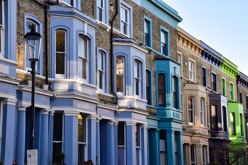 Photograph of a row of multi-story residential buildings with Victorian-style facades, featuring large arched windows and decorative moldings. The buildings are painted in pastel shades of blue, teal,