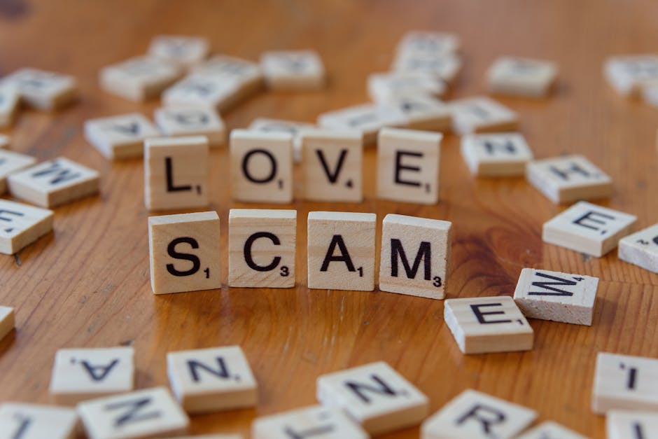 Close-up view of small wooden letter tiles arranged on a wooden surface, spelling out the words 'LOVE' and 'SCAM' in the center. The tiles are surrounded by scattered similar tiles with random letters