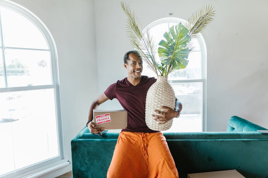 A man is standing inside a bright, well-lit room with large, arched windows that allow natural light to fill the space. He is smiling and holding multiple items in his hands: a cardboard box labeled '