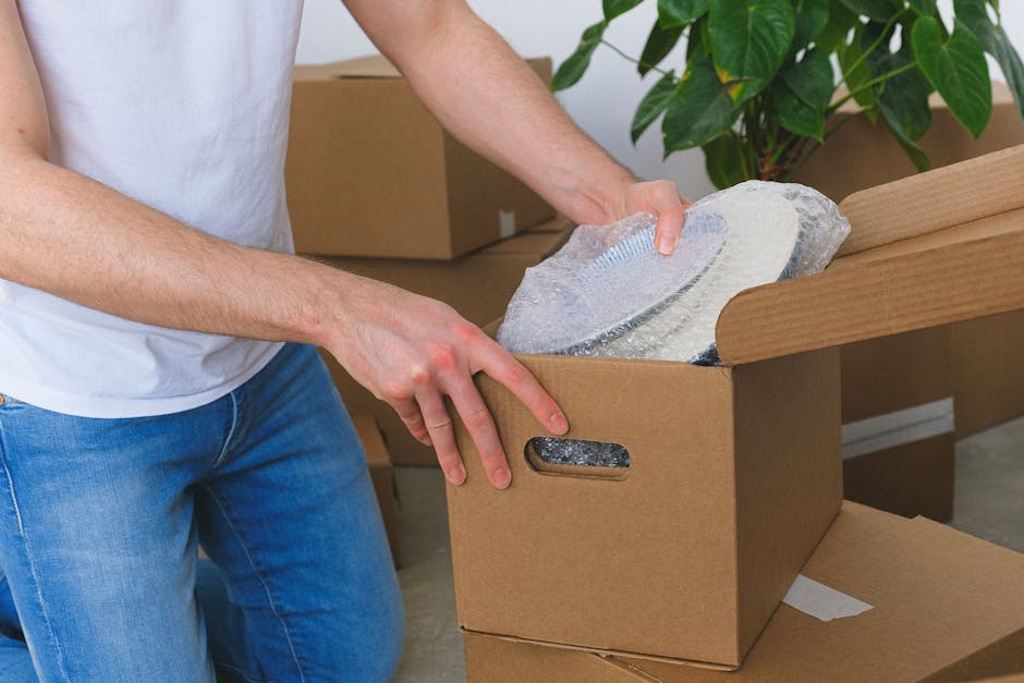 A man is standing inside a bright, well-lit room with large, arched windows that allow natural light to fill the space. He is smiling and holding multiple items in his hands: a cardboard box labeled '