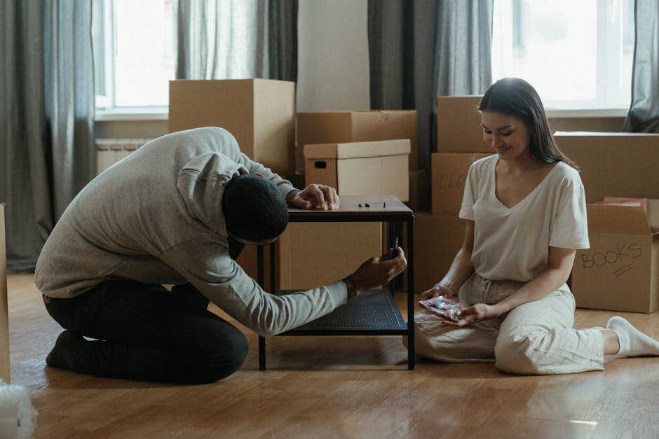 A young man and woman are indoors, surrounded by packed cardboard boxes, during the home relocation process. The woman is seated at a wooden table, using a screwdriver to secure or assemble items, whi